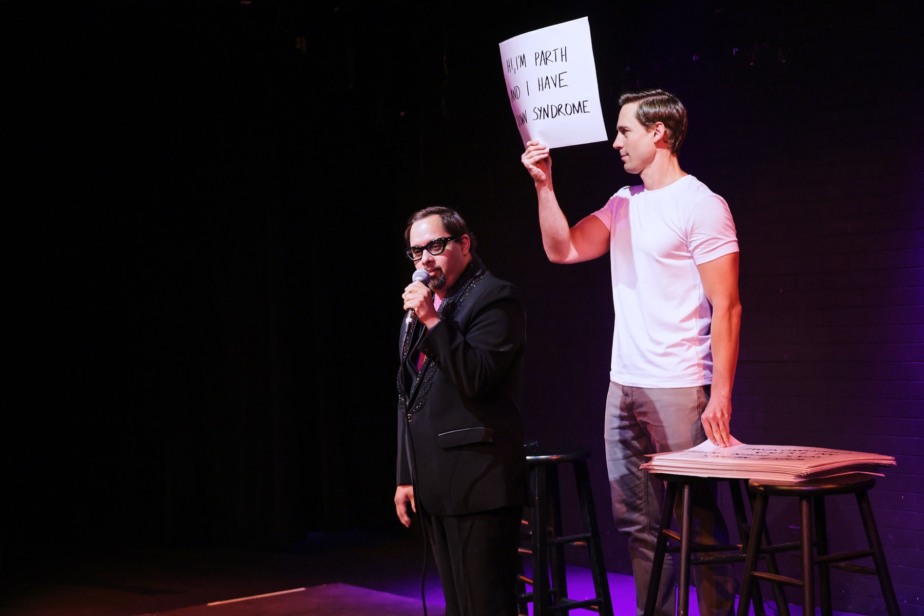 Parth Bhatt performing stand-up while Corey Martin Craig holds up a cue card that reads: Hi, I'm Parth and I have Down syndrome